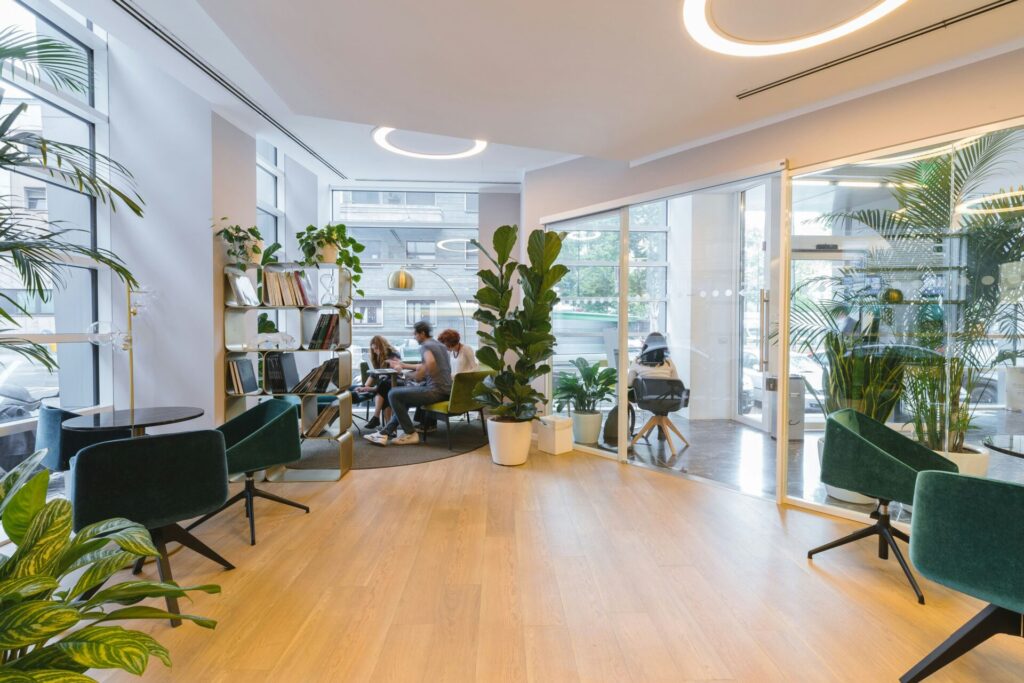 A modern office space with glass windows, lots of plants seating and people meeting